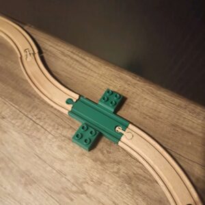 Carril verde impresso em 3D a unir pistas de madeira, exibindo abas laterais com encaixes para blocos de construção grandes.