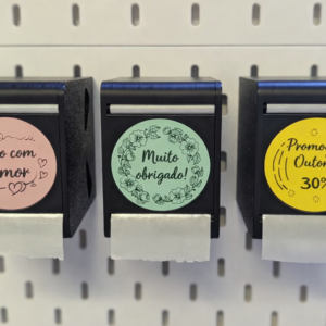 Três dispensadores de etiquetas pretos impressos em 3D no painel IKEA Skadis, com autocolantes informativos na frente.