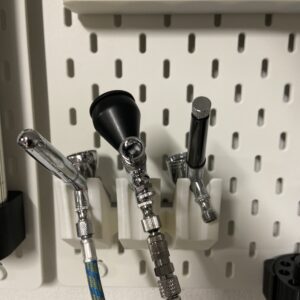 Três aerógrafos de precisão, um deles com mangueira ligada, organizados num suporte branco 3D no painel IKEA Skadis.