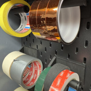 Vários rolos de fita adesiva e fita isoladora organizados num suporte horizontal preto no painel IKEA Skadis.