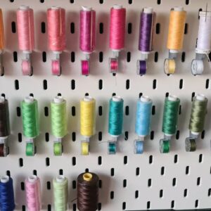 Painel IKEA Skadis organizado com suportes 3D para linhas de costura e canelas coloridas.