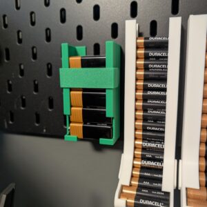 Painel IKEA Skadis com um suporte verde impresso em 3D para pilhas retangulares de 9V, ao lado de dispensadores brancos para pilhas AAA e AA.